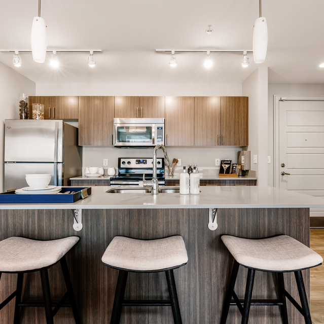 Cadence Union Station Apartments - Modern Kitchen with White Island, Bar Stools, Stainless Steel Appliances, and Wooden Cabinets