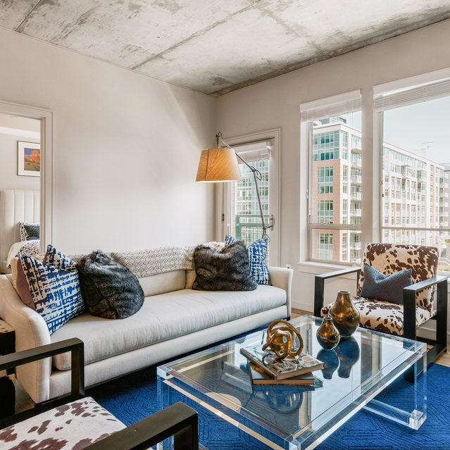 Cadence Union Station Apartments - ModernLliving Room with White Sofa, Blue Pillows, Glass Coffee Table, Black Armchairs, and Large Windows 